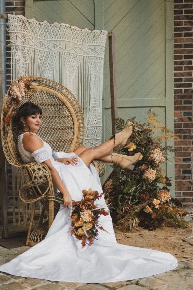 Femme en robe blanche assise sur une chaise, tenant un bouquet de fleurs, décor rustique.