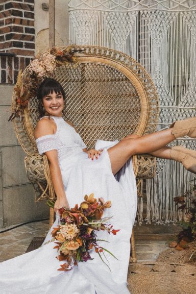 Femme en robe de mariée assise sur une chaise en osier, tenant un bouquet de fleurs.