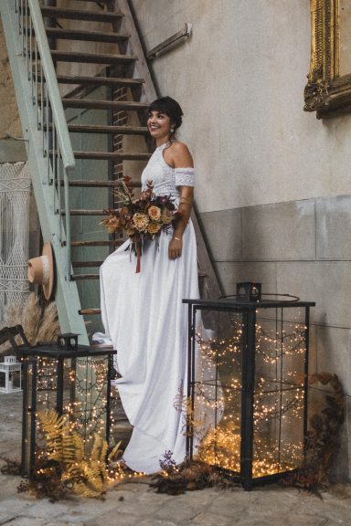 Femme en robe blanche portant un bouquet, debout près de lanternes dans un décor rustique.